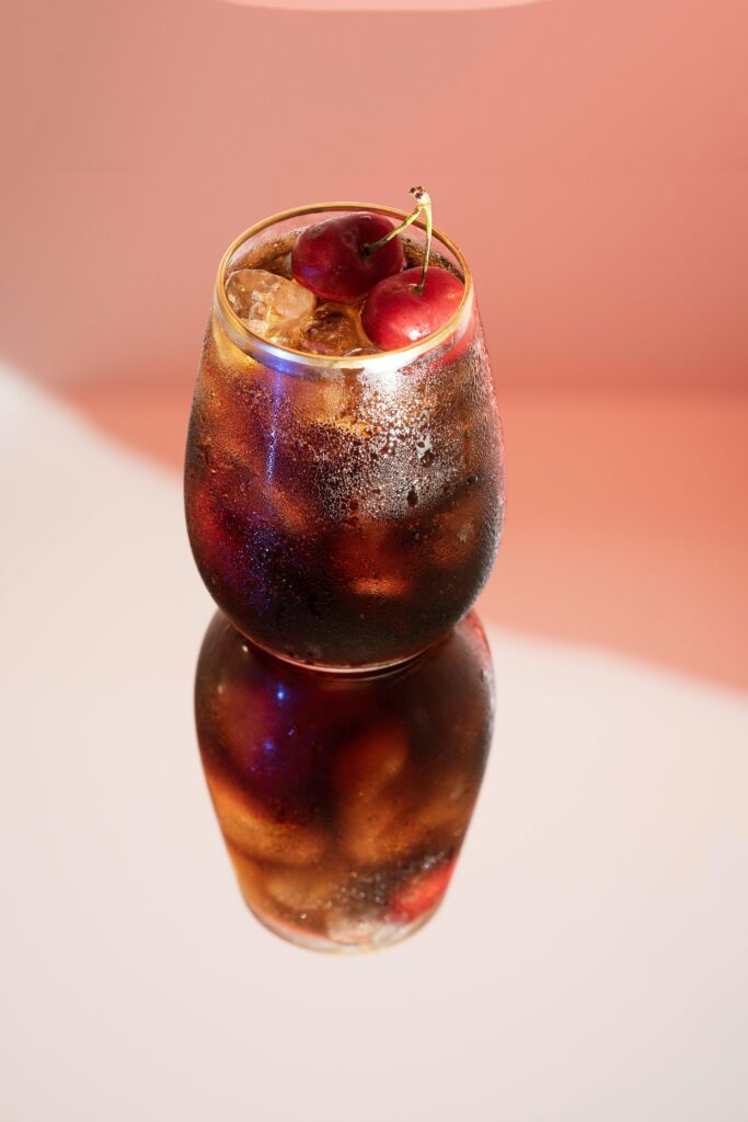 A cold glass of cherry cola with ice, featuring a vivid reflection and cherries on top.